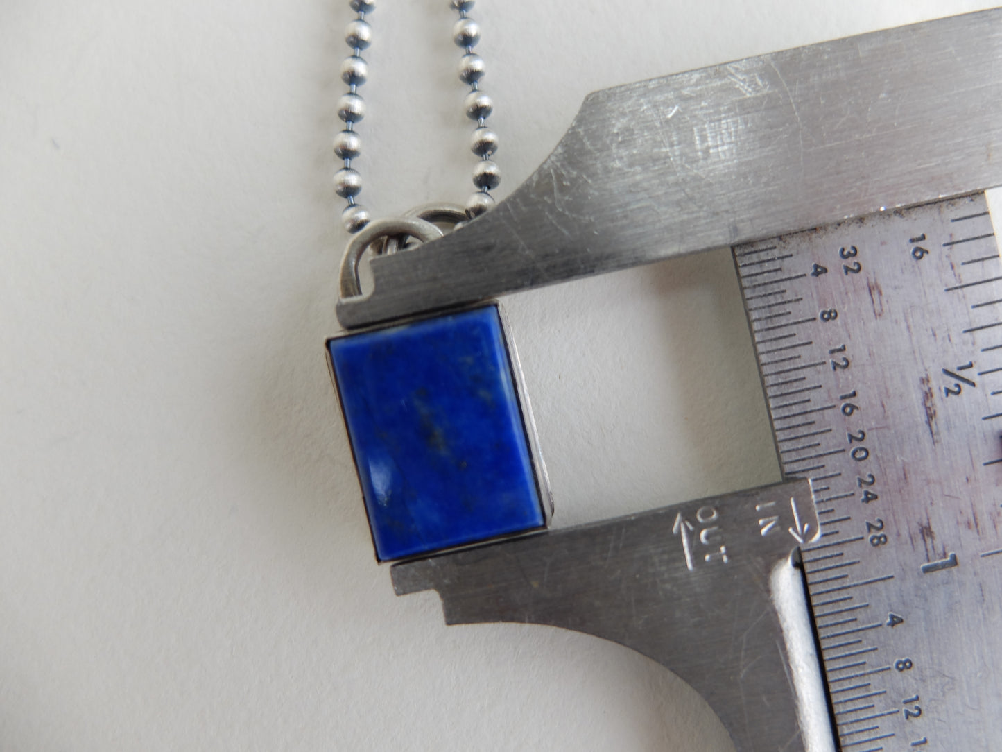Blue square pendant on a chain with a ruler for scale on a white background